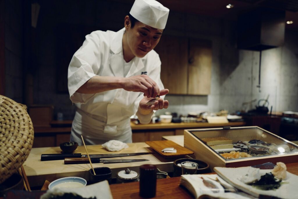 The image depicts a chef at an elegant sushi counter, passionately preparing seasonal ingredients to guests, enhancing their fine dining experience. The sophisticated ambiance and the chef's culinary expertise create a memorable culinary journey in this prestigious restaurant setting.