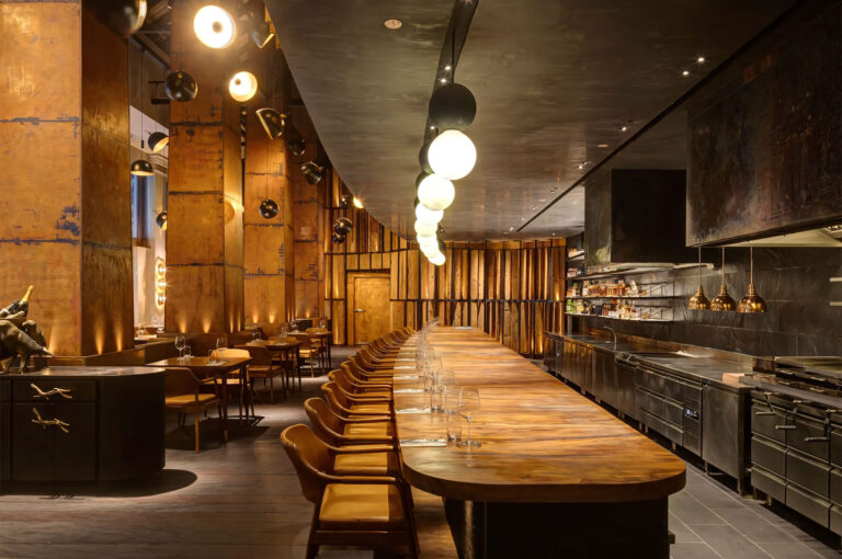 The modern, dimly lit interior of Burnt Ends restaurant, featuring a long wooden counter bar, yellow leather chairs, and dark, industrial-style walls and fixtures.