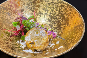 Gourmet Japanese appetizer, likely a savory roll or dumpling, topped with sea salt flakes and microgreens in a textured gold bowl.