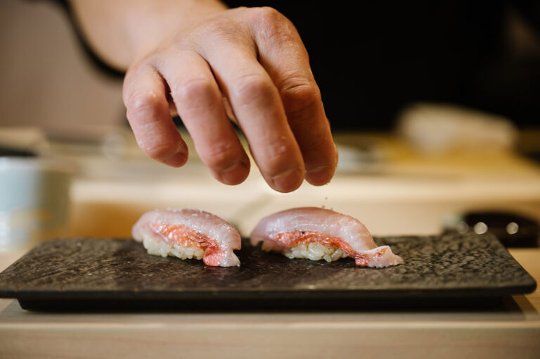 A sushi chef's hand delicately sprinkling salt or seasoning onto two pieces of pink and white nigiri sushi on a rectangular dark slate serving plate.