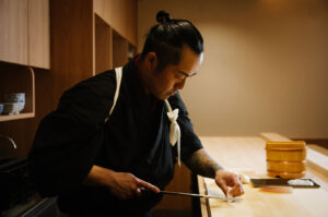 A sushi chef in a black kimono, with a visibly tattooed arm, using a long knife to precisely slice raw fish for sashimi on a wooden cutting board.