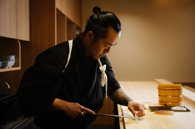 A sushi chef in a black kimono, with a visibly tattooed arm, using a long knife to precisely slice raw fish for sashimi on a wooden cutting board.