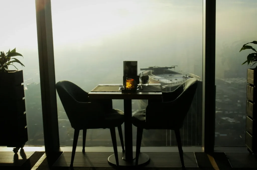 A window-side dining table for two with plush chairs overlooks a foggy cityscape. A candle and glasses are on the table, creating a cozy, intimate setting.