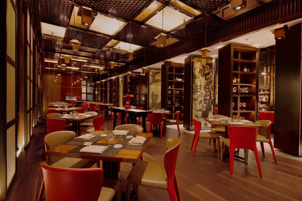 Elegant restaurant interior with warm lighting, wooden tables set with glassware, red and cream chairs, and decorative shelving, creating a cozy, inviting ambiance.