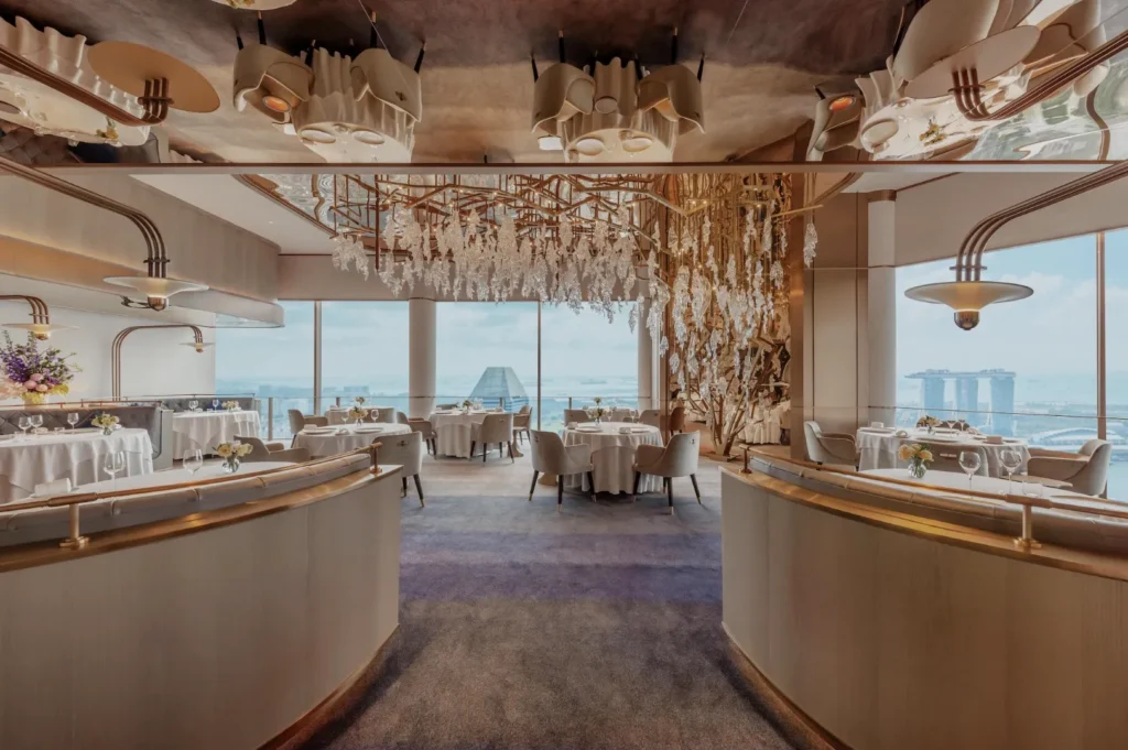 Luxurious restaurant interior with elegant chandeliers, beige decor, and floor-to-ceiling windows showcasing a panoramic city skyline view.