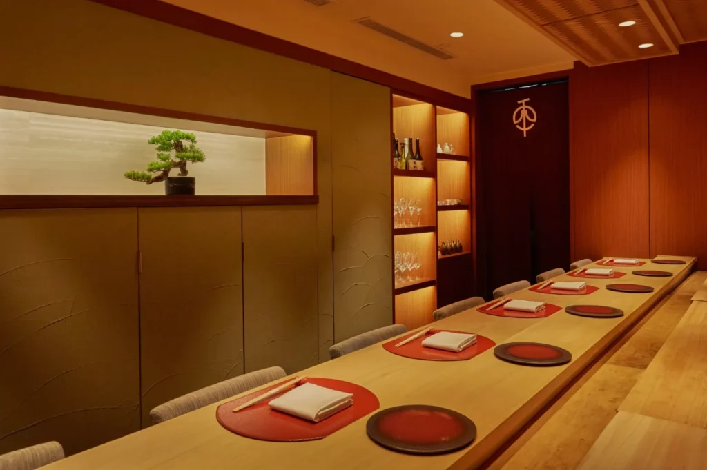 Elegant Japanese dining room with a long wooden table set for guests. Red placemats and chopsticks are neatly arranged. A bonsai tree and softly lit shelves add a serene touch.