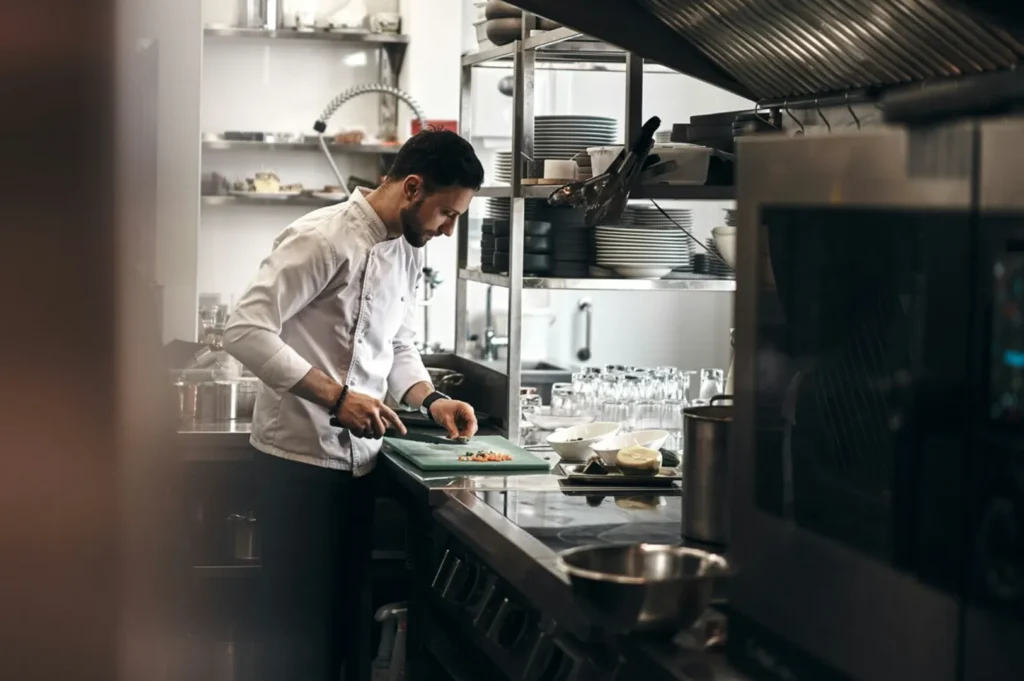 Chef in a white coat chopping vegetables on a cutting board in a modern, stainless-steel kitchen, conveying focus and professionalism.