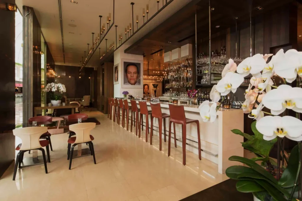 Chic, modern bar with high stools and plush seating, adorned with orchids and art. Warm lighting creates a sophisticated, inviting atmosphere.