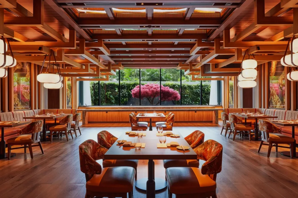 Warmly lit restaurant interior with wooden beams and tables set for dining. Large window reveals lush greenery, creating an inviting and elegant atmosphere.