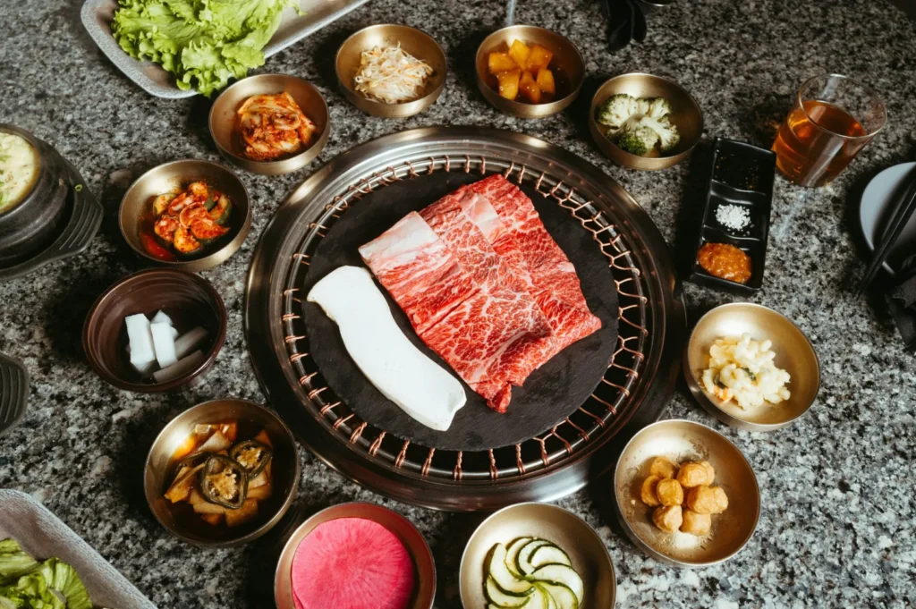 A Korean BBQ setup with marbled meat and a white strip on a grill, surrounded by leafy greens, pickles, kimchi, and various side dishes on a granite table.