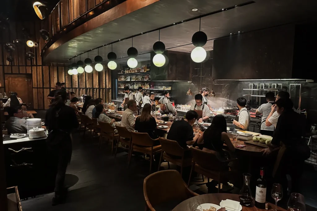 A dimly lit, bustling restaurant with chefs cooking behind a bar. Patrons seated on wooden chairs enjoy food under spherical pendant lights. Cozy ambiance.