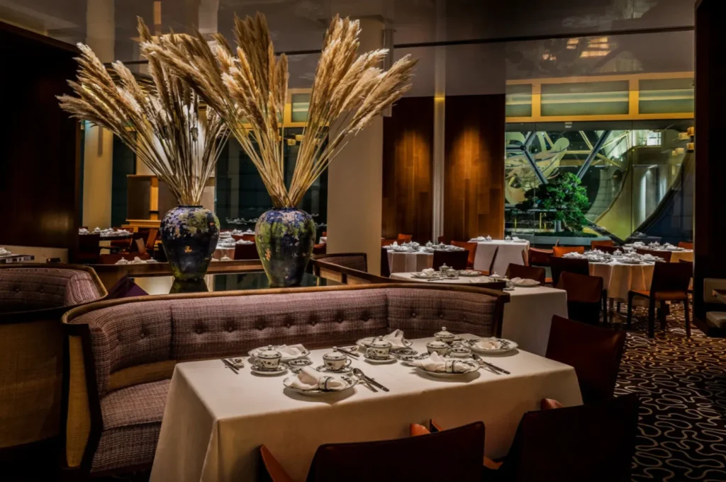 Elegant restaurant interior with neatly set tables, plush seating, and large vases of pampas grass. The ambiance is warm and sophisticated.