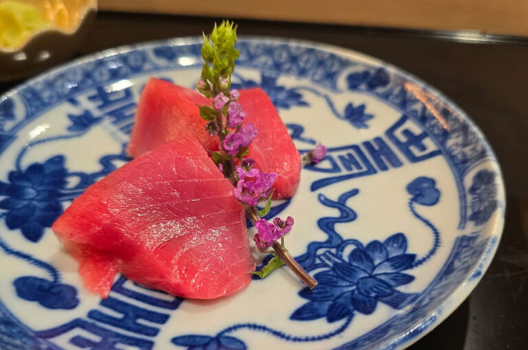 Three bright red pieces of maguro (tuna) sashimi beautifully plated on a white and blue Japanese ceramic dish, garnished with a sprig of purple edible flowers.