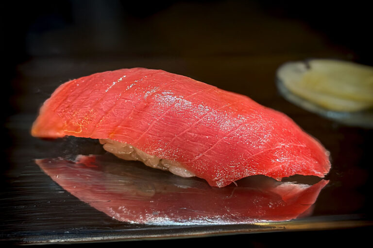 Macro shot of a single piece of vibrant red, finely scored otoro (fatty tuna) nigiri sushi, beautifully reflecting on a black serving plate.