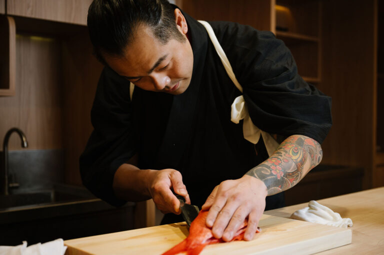 Close-up of a Japanese chef wearing a black uniform and white apron, slicing a piece of red snapper or red fish on a wooden cutting board with a knife.