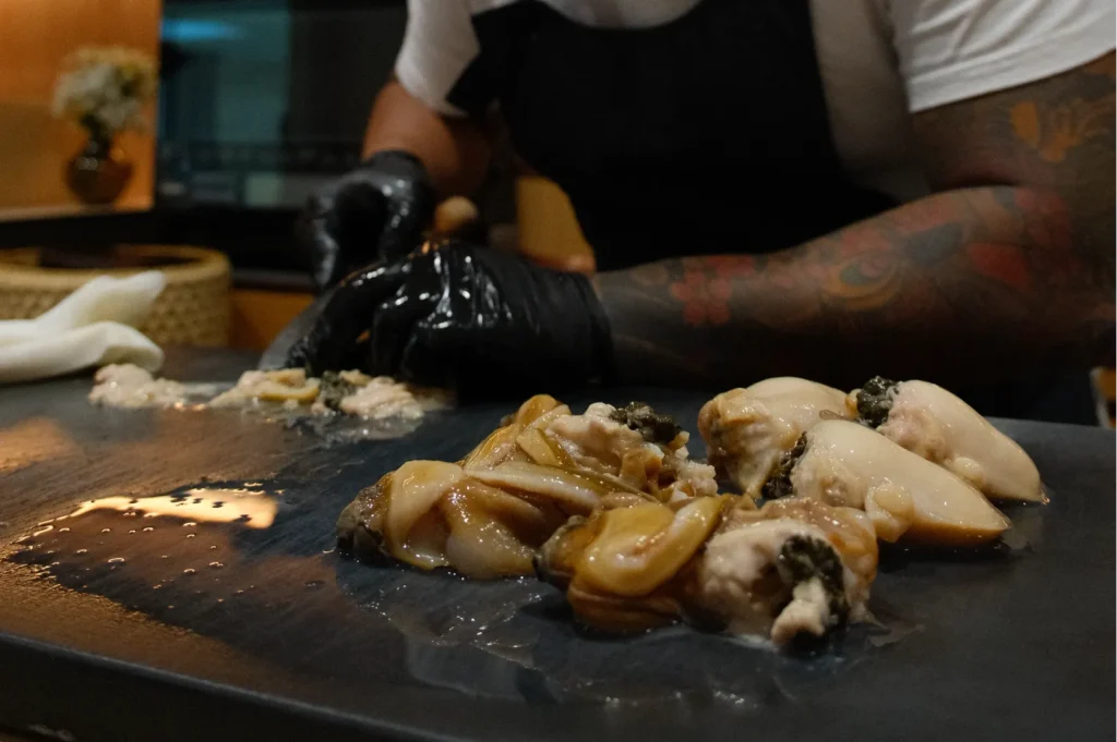 Chef Masa with tattooed arms and black gloves prepares raw clams on a dark countertop, creating a focused and professional culinary atmosphere.