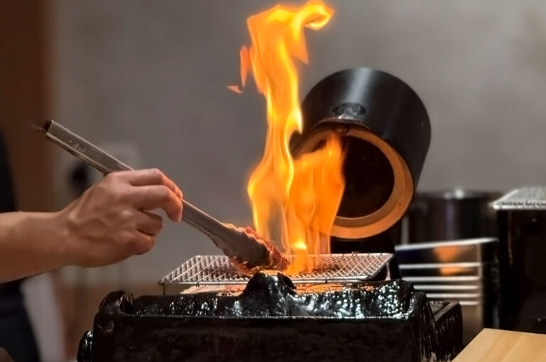 A chef's hand using tongs to cook or sear a piece of meat on a small grill, with a large, dramatic flame rising from the charcoal.