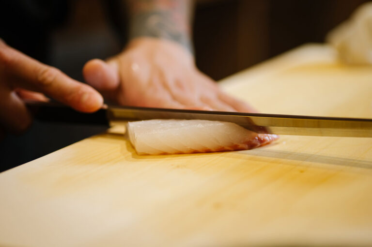 Extreme close-up of a chef's hand guiding a long, thin knife to perfectly slice a piece of white fish, likely hirame or flounder, for sashimi.