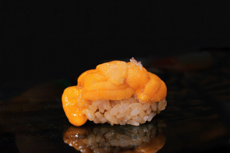 A single piece of rich, bright orange uni (sea urchin) nigiri sushi, served directly on rice on a dark surface.