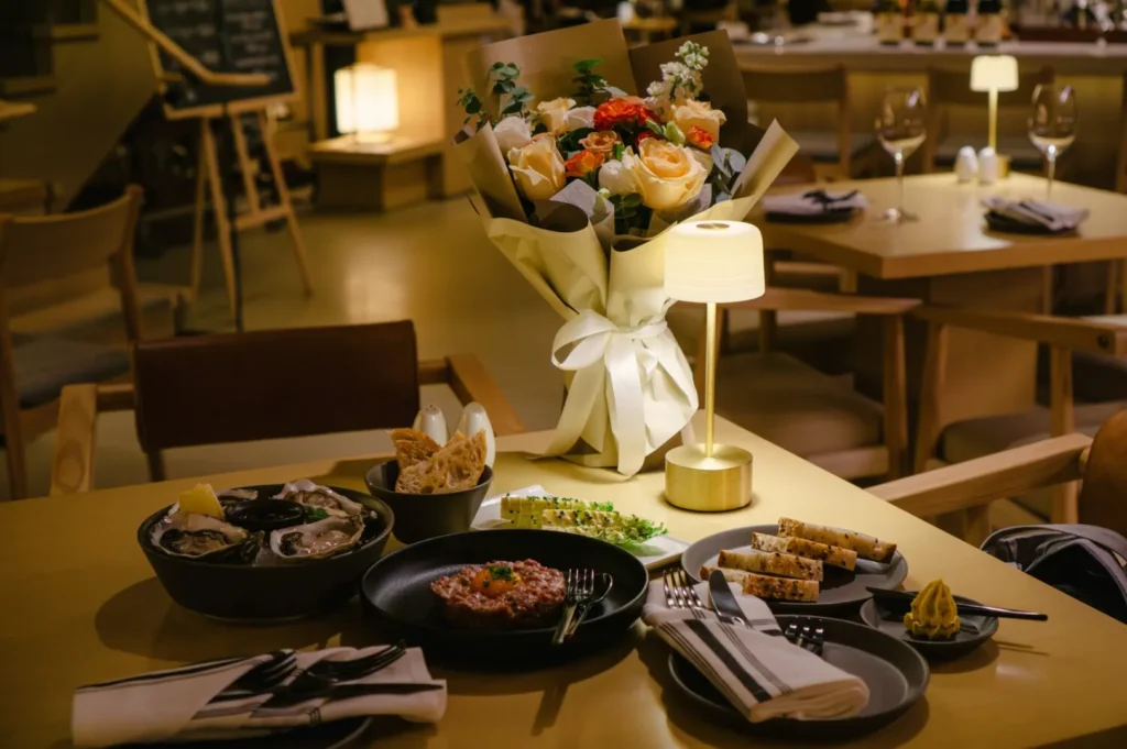 A warmly lit restaurant table set for dinner, featuring a bouquet of roses, elegant tableware, and a variety of dishes, creating a cozy atmosphere.