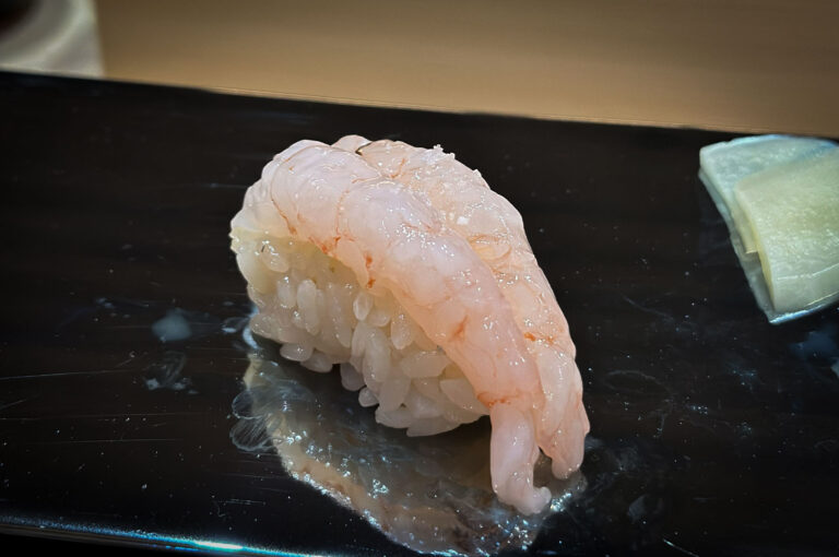 Close-up of a single piece of delicate, pale pink ama-ebi (sweet shrimp) nigiri sushi served on a dark plate, showing its reflection.