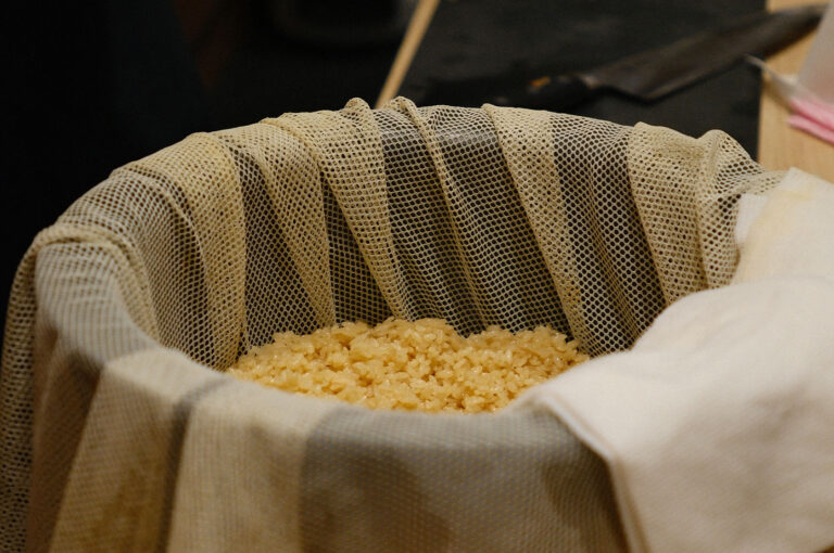 Close-up of perfectly cooked, seasoned, slightly yellow-tinged sushi rice resting in a wooden barrel lined with a mesh steaming cloth.