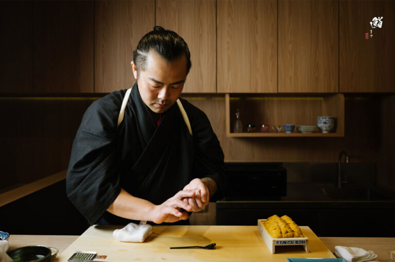 A sushi chef preparing food at a wooden counter, with a platter of fresh, bright yellow uni (sea urchin) in a box next to him.
