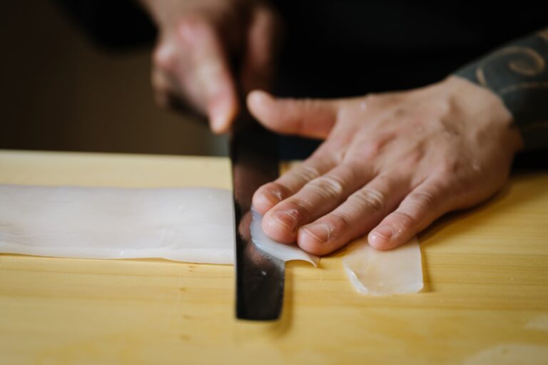 A skilled chef is expertly cutting fresh sashimi with a traditional Japanese knife on a wooden cutting board, showcasing the precision required in Japanese cuisine. The scene captures the artistry of preparing raw fish, a key component of dishes like nigiri sushi and sashimi, highlighting the freshness of the seafood.