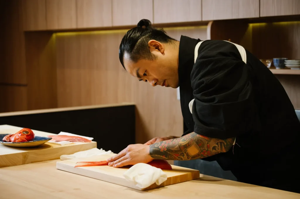 A focused Chef Masa with tattoos, in a black uniform, skillfully slices salmon in a modern kitchen. Plates of seafood and a wooden interior create a calm ambiance.
