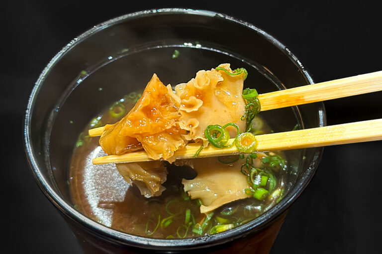 Two wooden chopsticks lifting a piece of cooked abalone or shellfish and wood ear mushroom from a small bowl of clear soup garnished with chopped green onions.