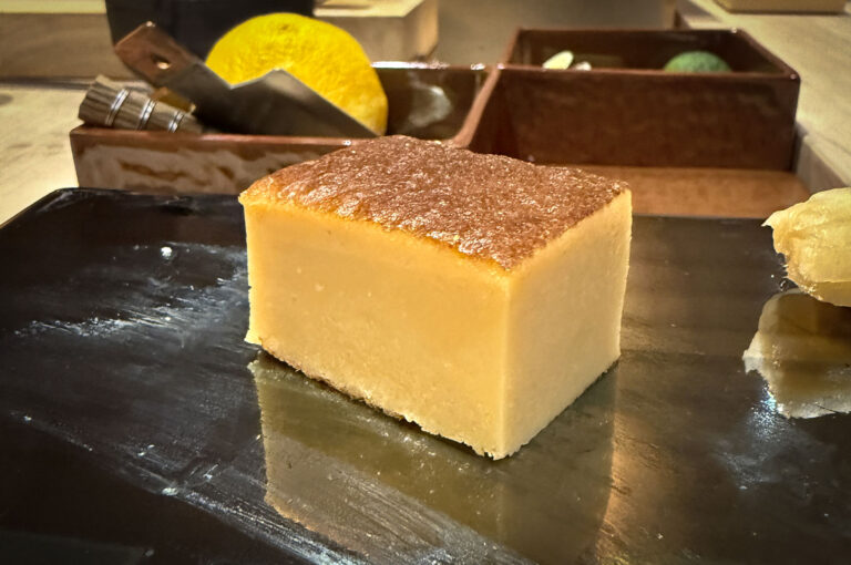 A square piece of golden-brown, rich custard or sponge cake, resembling a piece of thick Tamagoyaki or a Castella cake, served on a dark, reflective plate.