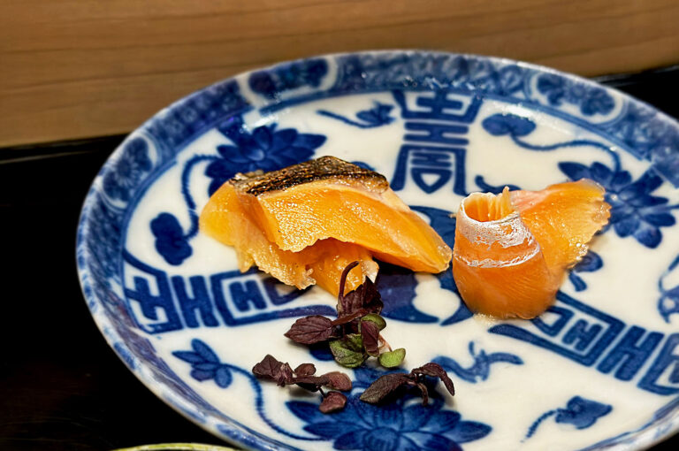 Two pieces of seared, orange salmon sashimi with crispy skin, delicately plated on a blue and white patterned ceramic dish next to dark microgreens.