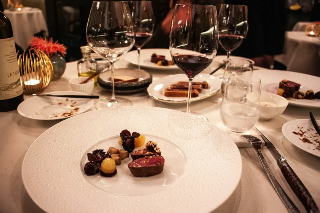 Close-up of a gourmet meal on a white textured plate, featuring rare meat topped with berries. The fine dining table includes several glasses of red wine and a lit candle.