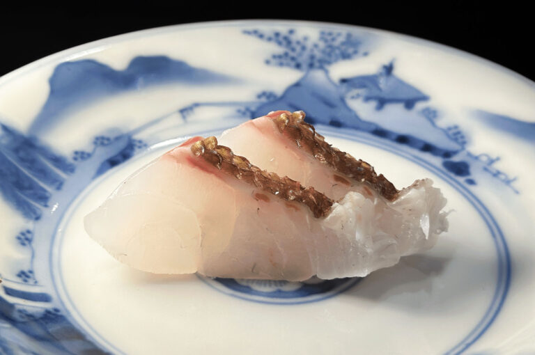 Two slices of light pink and white sashimi, likely Buri (Japanese amberjack) or a similar white fish, with brown seasoning on top, served on an antique-style blue and white ceramic plate.