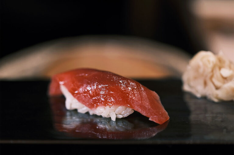Close-up of a single piece of glossy, red maguro (tuna) nigiri sushi resting on a dark, reflective plate with a piece of ginger in the background.