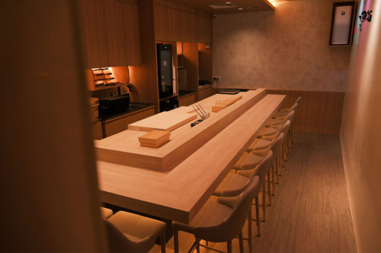 The intimate and minimalist interior of an omakase or sushi restaurant, featuring a long, light wooden counter bar with ten empty seats and warm lighting.