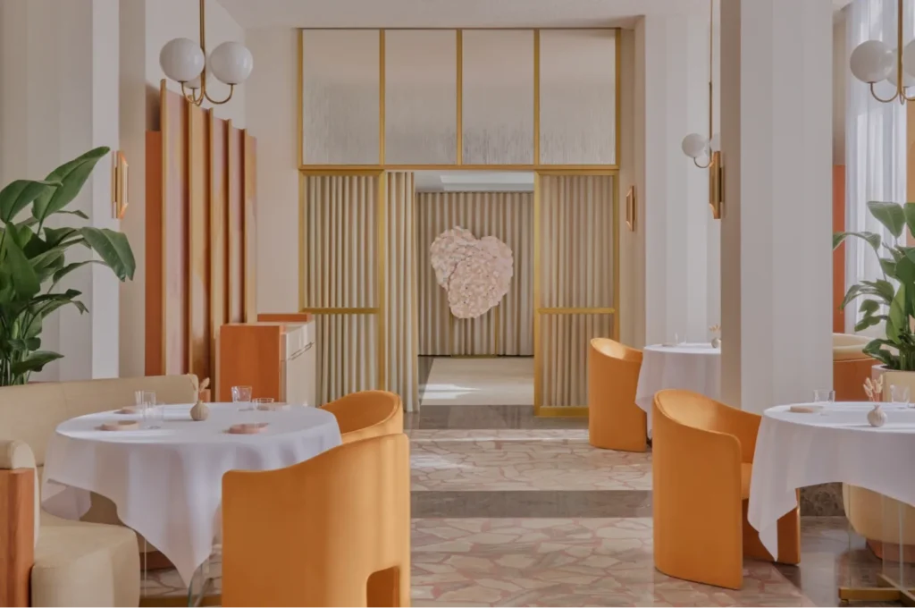 A modern restaurant dining room with white tablecloths, orange chairs, and a large heart-shaped floral art installation in the background.