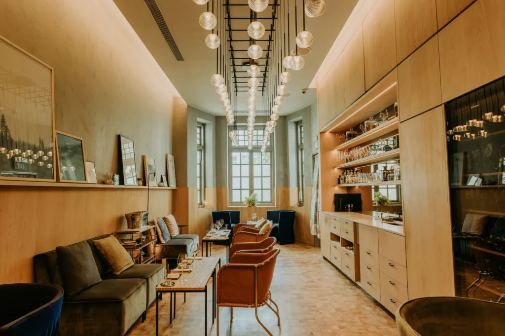 Stylish, ambient lounge with warm lighting, leather chairs, and a central long table. Art decorates the walls, and a bar with glassware appears on the right.