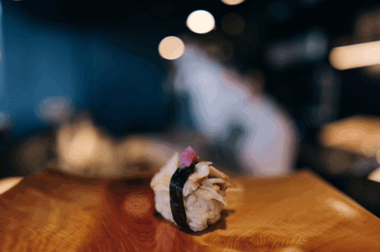 Single piece of shellfish sushi garnished with pink roe, resting on a wooden board against a softly blurred omakase backdrop. The shallow depth of field isolates the handcrafted bite, capturing the quiet reverence and visual poetry of chef’s choice dining.