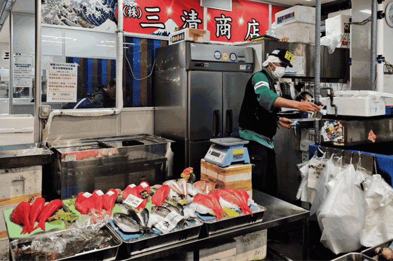 Interior of a traditional Japanese seafood shop with fresh seasonal fish and tuna cuts displayed on ice-lined green trays. A masked worker prepares seafood behind the counter, surrounded by styrofoam boxes and stainless equipment. The red company sign and iconic 'Great Wave' artwork evoke cultural pride and meticulous sourcing—hallmarks of authentic Japanese cuisine.