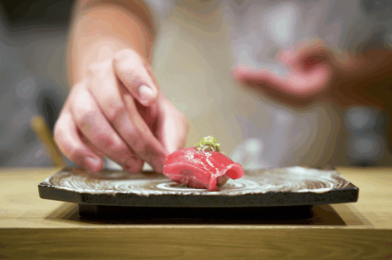 Close-up of a sushi chef placing a slice of raw tuna atop vinegared rice on a ceramic plate. The chef’s hands, captured mid-motion, reflect precision and reverence for tradition—embodying the artistry and seasonal sensitivity at the heart of authentic Japanese dining.