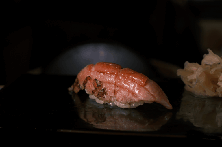 Glossy slice of fatty tuna (otoro) atop vinegared rice, placed on a dark reflective surface beside pickled ginger. The minimalist composition and rich texture evoke the purity, seasonality, and quiet luxury of authentic Japanese sushi craftsmanship.