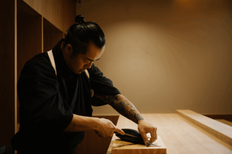 Chef in black attire with a tattooed arm expertly filleting fresh fish on a wooden cutting board inside Sushi Masa’s private dining room; the warm wood tones and subdued lighting frame the moment with quiet intensity, capturing the precision, artistry, and reverence behind each handcrafted dish in this intimate culinary setting.