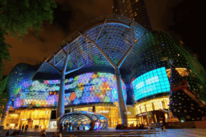 Nighttime view of ION Orchard in Singapore, its futuristic glass facade and illuminated canopy glowing with festive lights and luxury storefronts like Louis Vuitton and Cartier. A towering Christmas tree adds seasonal warmth to the upscale atmosphere. This iconic mall marks the gateway to Orchard Road’s sushi trail, where Japanese culinary artistry meets Singapore’s high-end dining culture.
