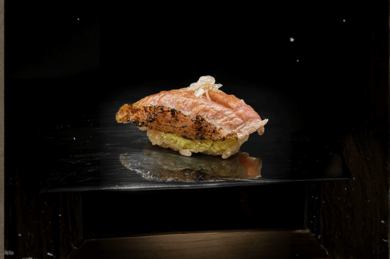 Elegant piece of seared sushi artfully plated on a reflective black surface, featuring charred tuna atop seasoned rice with green wasabi and delicate garnishes. The minimalist composition and dramatic lighting evoke the precision and beauty of high-end Japanese cuisine, capturing the essence of Orchard Road’s refined sushi experience.