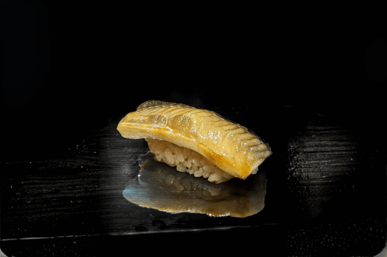 Glossy piece of eel sushi resting on a reflective black surface, its lacquered glaze catching the light above tightly packed rice. The minimalist presentation and rich texture evoke the depth and elegance of traditional Japanese flavors, spotlighting Orchard Road’s dedication to culinary craftsmanship.