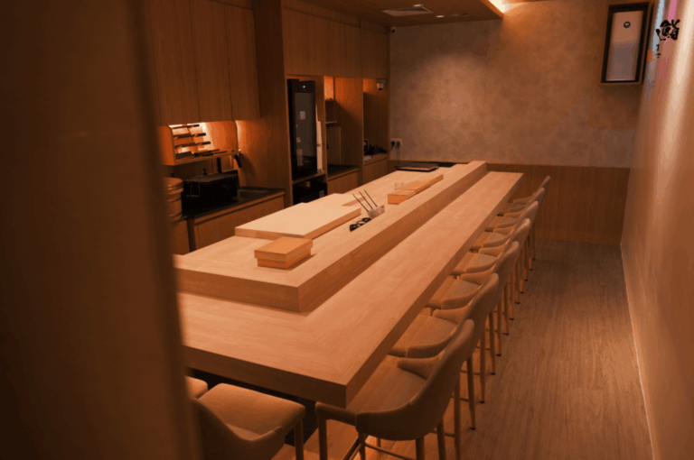 Minimalist omakase sushi counter with light wood finishes and cushioned seating for ten, set under warm ambient lighting. Wooden boards and chopsticks await guests, while the open kitchen hints at intimate chef-led service. This serene space reflects Orchard Road’s devotion to Japanese culinary precision and quiet luxury.
