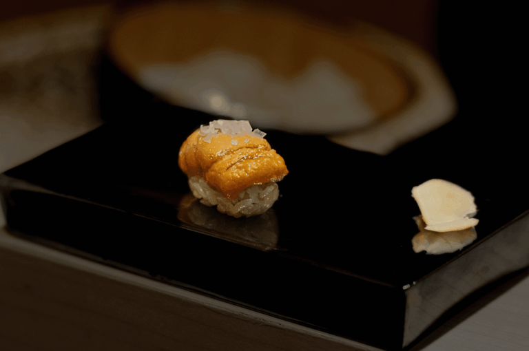 Luxurious uni nigiri presented on a glossy black plate, its vibrant orange sea urchin glistening with coarse salt atop seasoned rice. A sliver of pickled ginger rests beside it, while a wooden bowl in soft focus adds warmth to the minimalist composition. This dish captures the indulgent purity of Japan’s finest flavors, as celebrated in Orchard Road’s elite sushi scene.