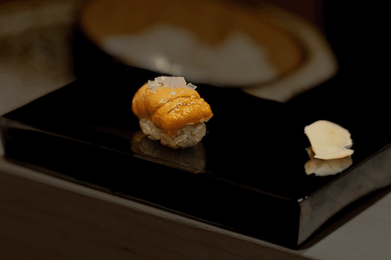 Golden-orange uni sushi garnished with finely chopped onions, plated on a glossy black dish at Sushi Masa by Ki-Setsu; minimalist presentation highlights the delicacy’s creamy texture and seasonal elegance.