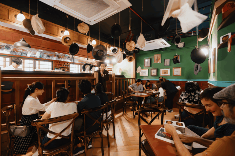 Interior of a cozy Singapore café offering a unique dining experience where guests dine amid eclectic decorations — hanging hats, framed art, and warm wooden textures. This multi-room venue blends sophisticated design with human connection, creating unforgettable memories through delicious food, desserts, and drinks. Diners explore familiar dishes and inspired cuisine from a curated menu, with cocktail pairing options and non-alcoholic drinks available. Located near Mount Faber and part of Southeast Asia’s best dining experience, this restaurant evokes the charm of the officer’s club and the wonderland upside concept. Whether booked for event Sat, event Mon, or event Fri, tables are served with care by staff behind the bar, completing a journey through taste, sight, and decor. Tickets sold are non-refundable and non-exchangeable, making this venue one of the best gifts for those seeking a multi-sensory dining experience in Singapore.